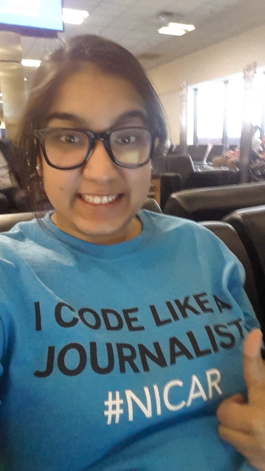 A blurry selfie of a young woman smiling in an airport lounge, wearing black-framed glasses and a bright blue t-shirt printed in bold with the words "I Code Like A Journalist #NICAR".